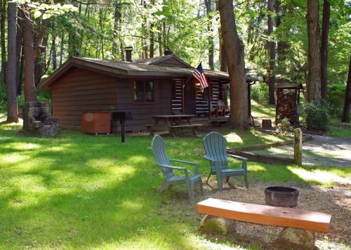 A cozy cabin in a wooded area, featuring a picnic table, chairs, and an American flag, surrounded by lush greenery.
