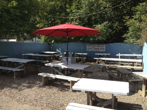 Outdoor picnic area with wooden tables and a red umbrella, surrounded by greenery and a blue fence.