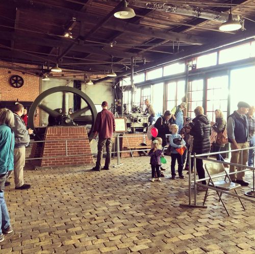 A crowd of people exploring a historic industrial exhibit with large machinery and brick walls.