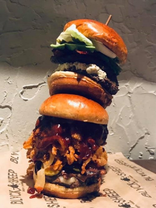 A towering stack of three gourmet burgers with various toppings, served on a paper-lined plate.