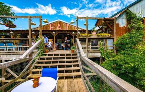 A rustic restaurant with a wooden deck, colorful chairs, and people dining under a blue sky with scattered clouds.