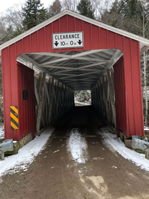 A red covered bridge with a clearance sign, leading into a snowy, tree-lined path.