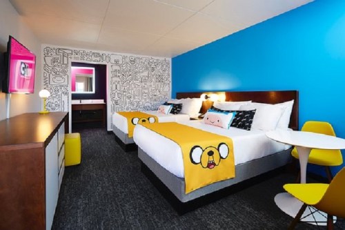A colorful hotel room featuring two beds with playful yellow blankets, a blue accent wall, and modern furniture.
