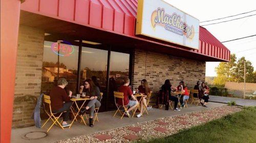 A cozy restaurant with outdoor seating, where people enjoy meals at small tables under a colorful sign that says "MacCheesy."