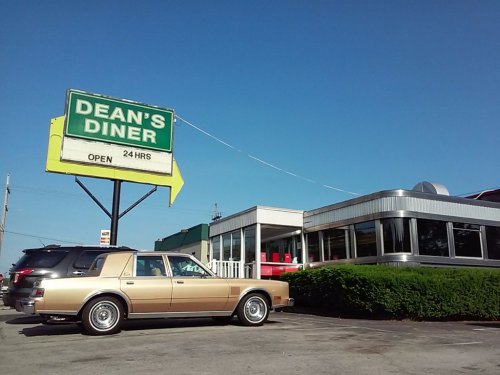 A classic diner named Dean's Diner with a 24-hour sign and a vintage car parked outside under a clear blue sky.