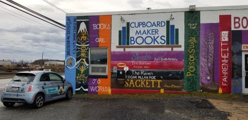 Colorful mural on a bookstore wall featuring book titles and artistic designs, with a small car parked nearby.