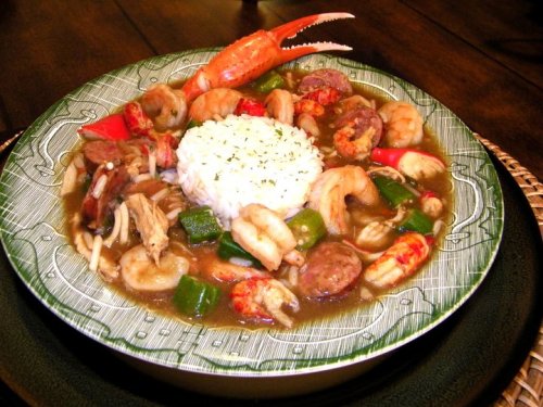 A colorful plate of gumbo with shrimp, sausage, vegetables, and a mound of rice, garnished with herbs.