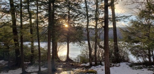 Sunlight filters through trees by a frozen lake, with patches of snow on the ground and a serene landscape in the background.