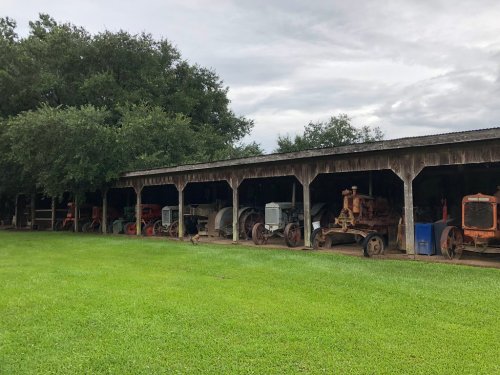 A row of vintage tractors under a wooden shelter, surrounded by green grass and trees, on a cloudy day.