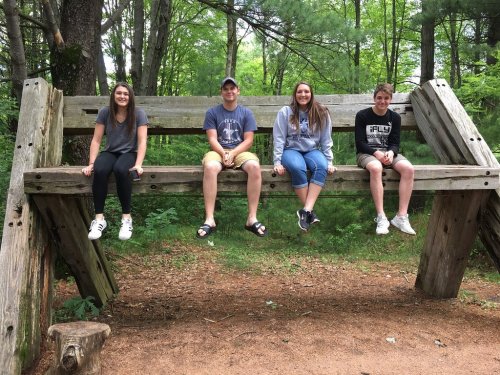 Four friends sit on a large wooden swing in a forested area, smiling and enjoying their time together.