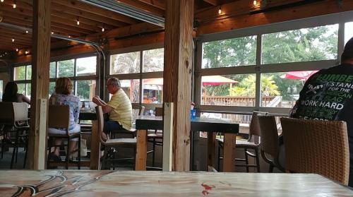 A casual dining space with wooden beams, patrons at tables, and large windows overlooking a patio with umbrellas.