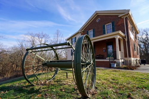 A vintage green cart wheel lies on the grass in front of a brick house with green shutters and a clear blue sky.