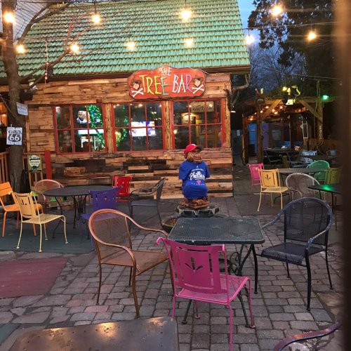 A person sits alone at a colorful outdoor bar with string lights, surrounded by empty chairs and a rustic wooden structure.