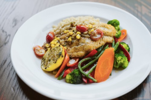 A plate of grilled fish with rice, mixed vegetables, and a lemon slice, garnished with cherry tomatoes and corn.