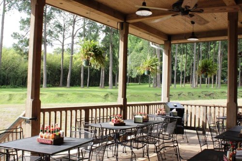 A spacious outdoor dining area with tables, a grill, and hanging plants, surrounded by trees and greenery.