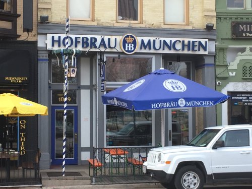Exterior of Hofbräu München restaurant with blue umbrellas and a white vehicle parked outside.