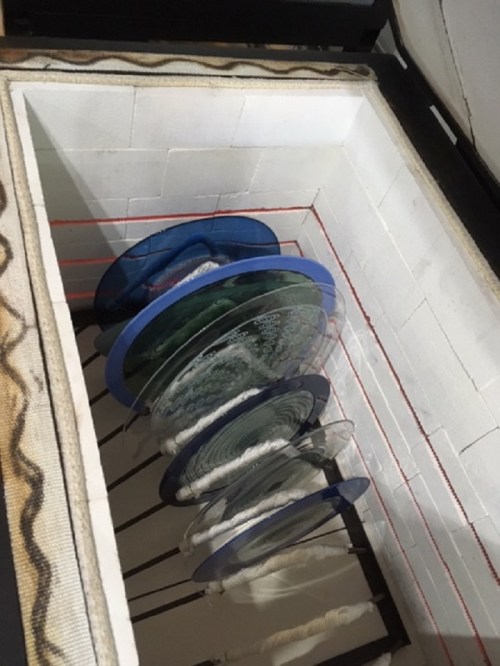 A top-down view of a kiln with stacked glass plates inside, showcasing various colors and patterns.