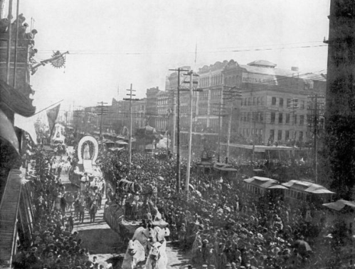 A historic parade with crowds lining the streets, featuring floats and buildings in the background.