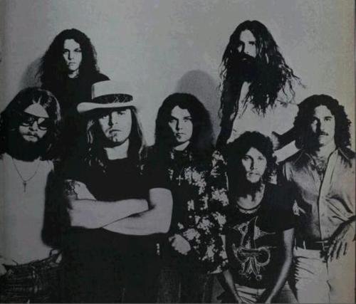 A group of seven men with long hair, dressed in 1970s fashion, posing together in a black and white photo.