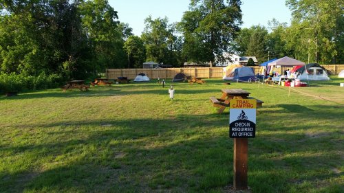 A grassy camping area with tents, picnic tables, and a sign indicating trail camping check-in at the office.