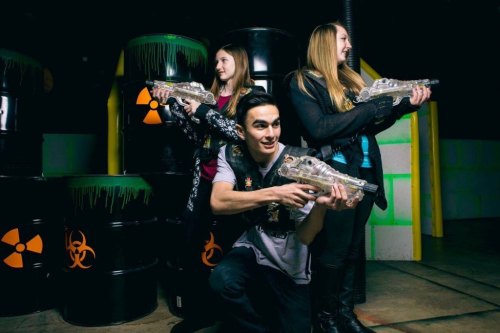 Three people pose with laser guns in a neon-lit indoor arena, with barrels in the background.
