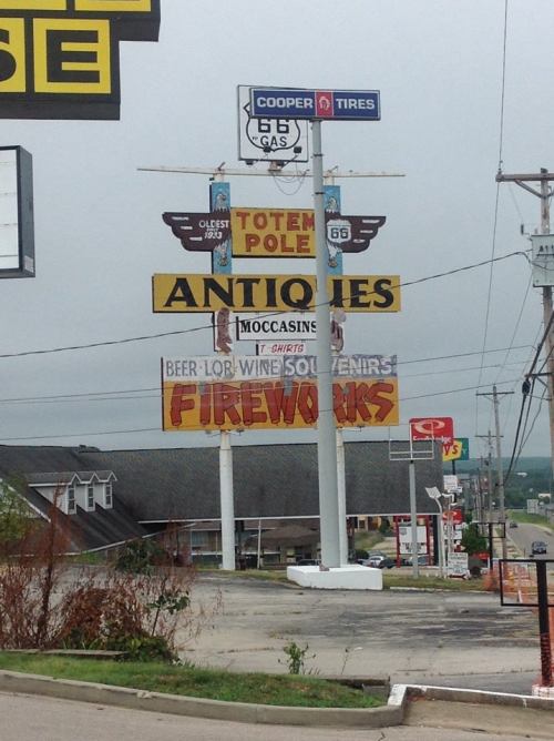 Signpost featuring various advertisements, including antiques, fireworks, and a gas station, under a cloudy sky.