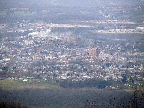 A panoramic view of a small town surrounded by hills, with buildings, smoke rising, and a cloudy sky.