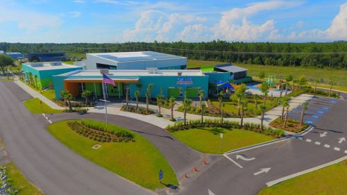 Aerial view of a modern building with colorful exterior, surrounded by palm trees and a clear blue sky.