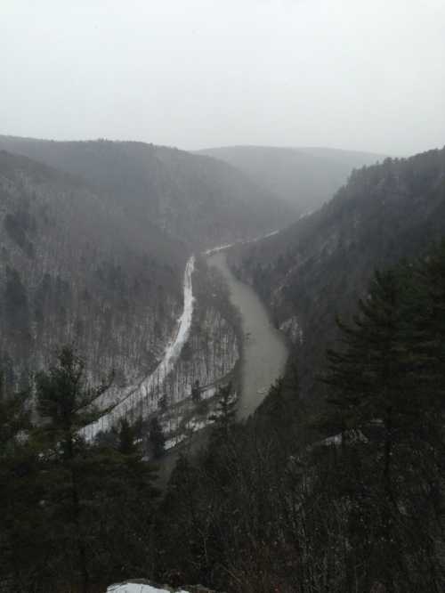 A misty landscape featuring a winding river surrounded by snow-covered hills and trees.