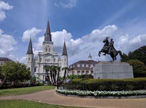 A historic church with spires and a statue of a horseman in a park under a blue sky with clouds.