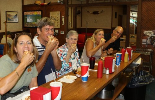 Five people sitting at a counter, enjoying hot dogs and drinks in a casual diner setting.