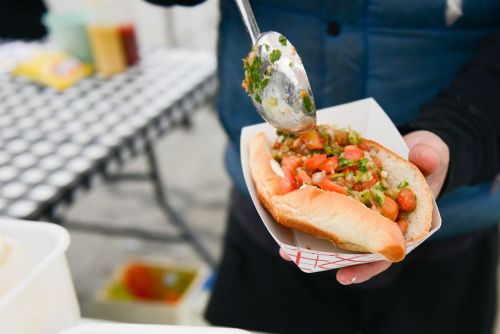 A person holds a hot dog topped with diced tomatoes and green onions, with a spoon in hand for serving.