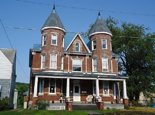 Victorian-style brick house with two towers, a porch, and a blue sky in the background.