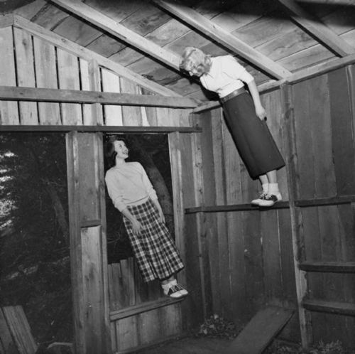 Two women in vintage clothing inside a rustic wooden structure, one standing on a ledge and the other leaning out of a window.