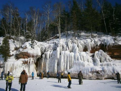 People exploring a snowy landscape with large ice formations and a forested hillside in the background.