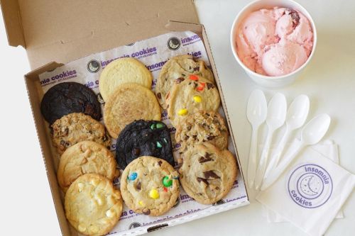 A box of assorted cookies from Insomnia Cookies, alongside a cup of pink ice cream and plastic spoons.