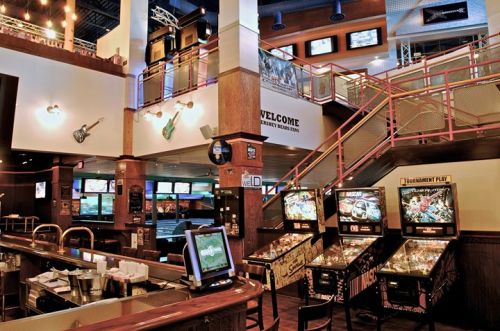 A lively bar with a multi-level layout, featuring pinball machines, screens, and a welcoming atmosphere.