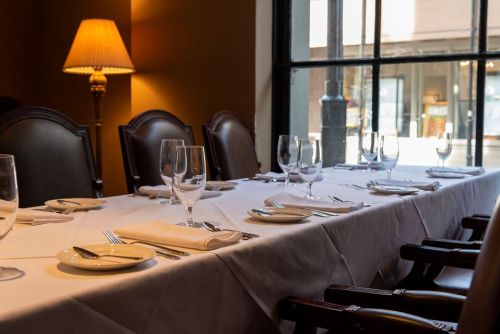 A elegantly set dining table with glassware and cutlery, illuminated by a warm lamp, near a large window.