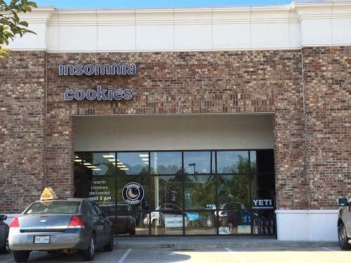 Exterior of Insomnia Cookies store with a brick facade, large windows, and a sign indicating late-night cookie delivery.
