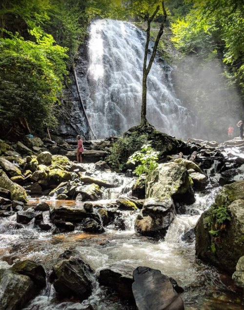 A waterfall cascades down rocky terrain, surrounded by lush greenery and mist, with a person standing nearby.