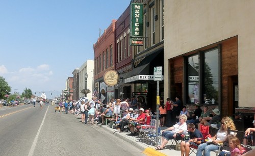 A busy street scene with people sitting in chairs along the sidewalk, waiting for an event in a small town.