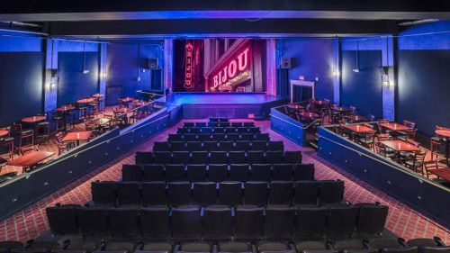 Interior of a theater with rows of seats, tables, and a stage featuring a "Bijou" sign in vibrant lighting.