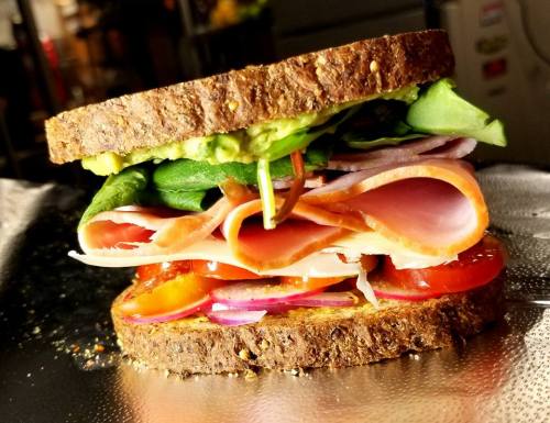A close-up of a sandwich with layers of ham, turkey, spinach, tomatoes, and avocado on whole grain bread.