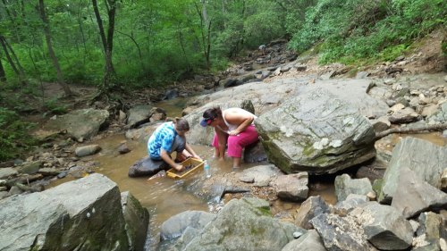 Two people are sitting by a stream, one using a pan to sift through rocks while the other observes nearby.