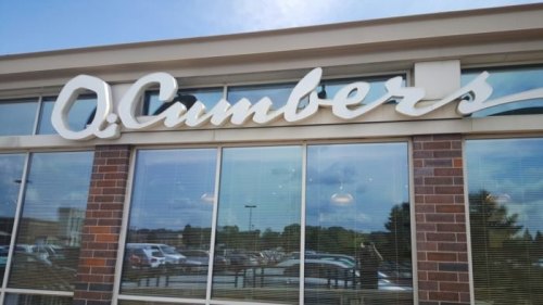 Sign for Q.Cumbers restaurant displayed on a building with large windows reflecting the sky and surrounding area.