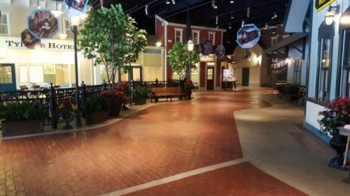 A charming indoor street with brick pathways, benches, and decorative plants, resembling a small town square.