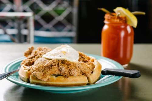 A plate with fried chicken and a fried egg on waffles, alongside a cocktail in a jar with a lemon slice.