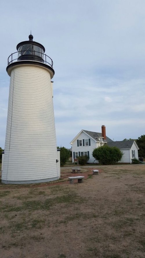 A white lighthouse stands next to a small house, surrounded by grassy land under a cloudy sky.