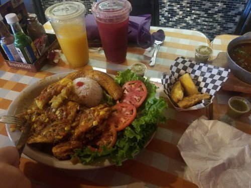 A colorful plate of grilled chicken with rice, salad, and plantains, accompanied by drinks and a side of wings.