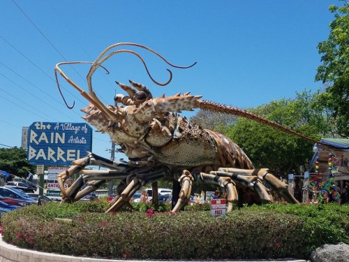 A large lobster sculpture outside the Rain Barrel Village of Artists, surrounded by greenery and colorful signs.